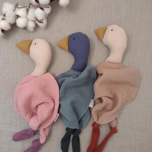 K&ouml;nnte beinhalten: Drei weiche G&auml;nse-Pl&uuml;schtiere in Pastellfarben. Jede Gans hat einen unterschiedlich farbigen Kopf und K&ouml;rperwickel: rosa, blau und hellbraun. Die Schn&auml;bel sind gelb und die Beine sind aus geknotetem Stoff in passenden Farben. Baumwollzweige sind im Hintergrund.