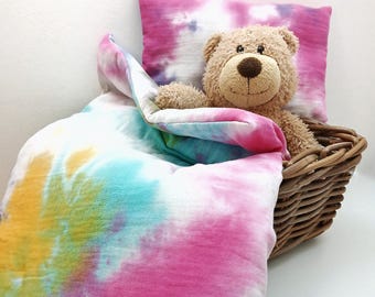 May include: A tie-dye patterned bedding set with a teddy bear resting on a pillow. The bedding is in shades of pink, blue, yellow, and white.