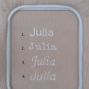 May include: A light beige fabric held in a white rectangular embroidery hoop. The fabric features four lines of white embroidered text, each reading the name Julia in a different font style, numbered 1 through 4 on the left side of the names.