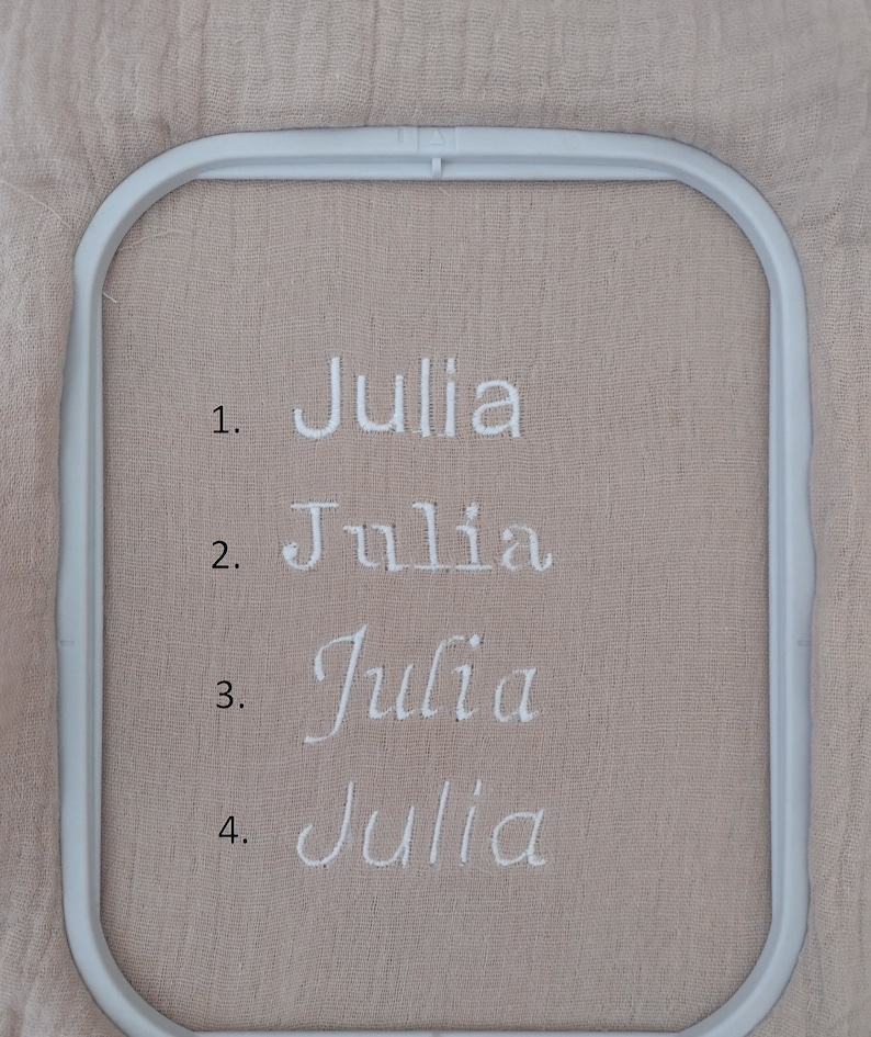 May include: A light beige fabric held in a white rectangular embroidery hoop. The fabric features four lines of white embroidered text, each reading the name Julia in a different font style, numbered 1 through 4 on the left side of the names.