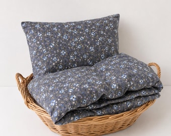 May include: A rectangular pillow and a folded blanket featuring a dark gray fabric with a repeating pattern of small light blue and white flowers. The bedding set is contained within a round, light brown woven wicker basket with two handles.