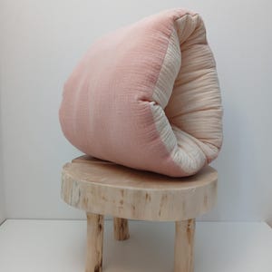 May include: A soft, folded cushion featuring a dusty pink exterior and a cream-coloured interior, resting on a small, round, light-coloured wooden stool with three legs. The fabric appears to be a textured, layered cotton muslin material.