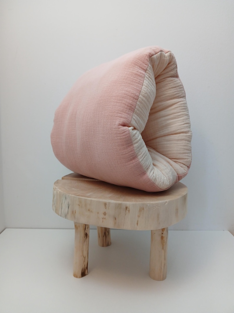 May include: A soft, folded cushion featuring a dusty pink exterior and a cream-coloured interior, resting on a small, round, light-coloured wooden stool with three legs. The fabric appears to be a textured, layered cotton muslin material.