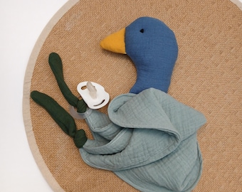 May include: A blue duck-shaped baby comforter with a yellow beak and a white pacifier. The comforter is on a round, woven mat with a light beige border. The duck has green legs.