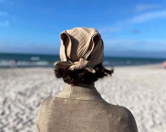 May include: A beige textured headscarf tied at the back with a knot, featuring two upright ear-like points at the top. The headscarf is worn with a matching beige knit sweater that has a ribbed collar and a textured, woven fabric construction.