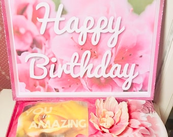 Może przedstawiać: Różowe pudełko prezentowe z brokatem z kwiatowym wzorem i napisem "Happy Birthday". W środku szalik, karta z napisem "You are AMAZING" i dekoracyjny kwiat.