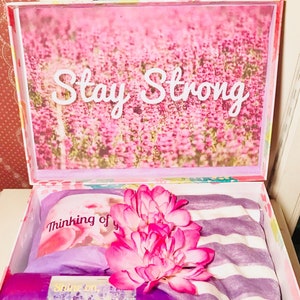 May include: A floral gift box with a pink and purple theme. The box contains a striped blanket, a rolled item, and a card that says "Thinking of you." The box lid has the words "Stay Strong" over a floral background.