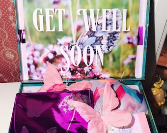 Może przedstawiać: Turkusowe pudełko prezentowe z kwiatowym wzorem i napisem "Get Well Soon" na pokrywie. Pudełko jest otwarte i zawiera fioletową torbę prezentową z wzorami motyli, różowego motyla i różowo-biały wzorzysty szalik.