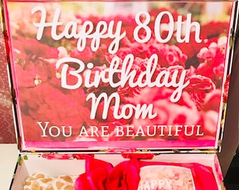 Może przedstawiać: Pudełko prezentowe z kwiatowym wzorem, zawierające szalik, kartkę i dekoracyjne kwiaty. Na pokrywie pudełka widnieje napis "Happy 80th Birthday Mom, You Are Beautiful". Szalik ma beżowo-biały wzór.