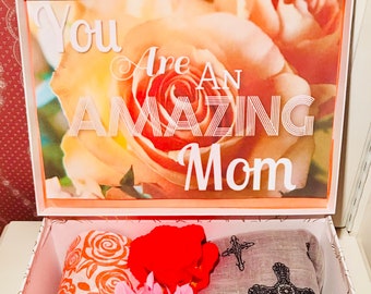 Może przedstawiać: Pudełko prezentowe z napisem "You Are An Amazing Mom" nad kwiatowym obrazem. W środku zwinięte szaliki z wzorami róż i krzyży, różowy kwiat i pomarańczowy przedmiot. Pudełko jest białe ze złotym kwiatowym wzorem.