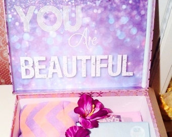 Może przedstawiać: Ozdobne pudełko z pokrywą z białym napisem "YOU Are BEAUTIFUL". Wewnątrz widoczny koc w zygzaki, karta z ilustracją motyla i fioletowe kwiaty.