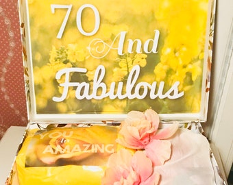 Może przedstawiać: Ozdobne pudełko na prezent z kwiatowym wzorem i napisem "70 And Fabulous". Pudełko zawiera żółtą bibułę, zapakowany prezent i różowe kwiaty. Pudełko jest ozdobione wizerunkami ptaków i kwiatów.