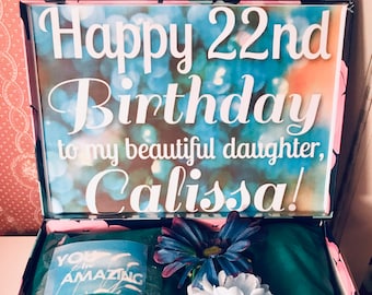Może przedstawiać: Niebieskie pudełko prezentowe z kwiatowym wzorem i różową pokrywką. Pokrywka jest otwarta i ma przezroczysty napis "Happy 22nd Birthday to my beautiful daughter, Calissa!" Wewnątrz pudełka znajduje się niebiesko-biały wzorzysty szal, niebieski kwiat, biały kwiat i niebiesko-biała torba prezentowa z napisem "You are amazing" na niej.
