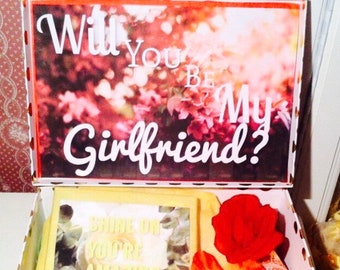 Może przedstawiać: Białe pudełko w złote kropki z czerwonym i żółtym kwiatem w środku. Pokrywa pudełka ma kwiatowy wzór z napisem "Will you be my girlfriend?" białymi literami.