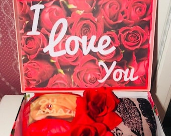Może przedstawiać: Pudełko prezentowe z pokrywą z motywem czerwonych róż i napisem "I Love You". Wewnątrz widoczny szalik w panterkę, czerwony przedmiot w kształcie flaminga i inne prezenty. Pudełko jest ozdobione wzorami kwiatowymi i paskami.