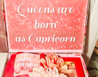 Może przedstawiać: Różowe pudełko prezentowe z kwiatowym wzorem i tekstem "Queens are born as Capricorn". Pudełko jest otwarte i zawiera beżowy koc z wzorem krzyża, czerwony worek prezentowy z tekstem "Happy Birthday" i dwa różowe kwiaty.