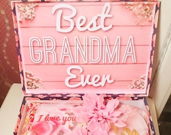Może przedstawiać: Różowe pudełko prezentowe z kwiatowym wzorem i pokrywką z napisem "Best Grandma Ever". Pudełko jest otwarte i zawiera poduszkę z różowo-białym wzorem kwiatowym, różowo-biały szal z kwiatowym wzorem i dwa różowe kwiaty.