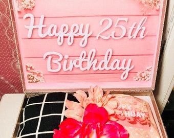 Może przedstawiać: Pudełko prezentowe na urodziny z różową pokrywą i białym napisem "Happy 25th Birthday". W środku czarno-biały kratkowany przedmiot, różowy przedmiot i zwinięty przedmiot z napisem "AMAZING". Na wierzchu duży czerwony kwiat.