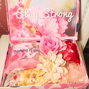 May include: A pink floral gift box with the words "Stay Strong" on the lid. Inside are pink and floral items, including a scarf, a flower, and a card that says "You are Amazing".