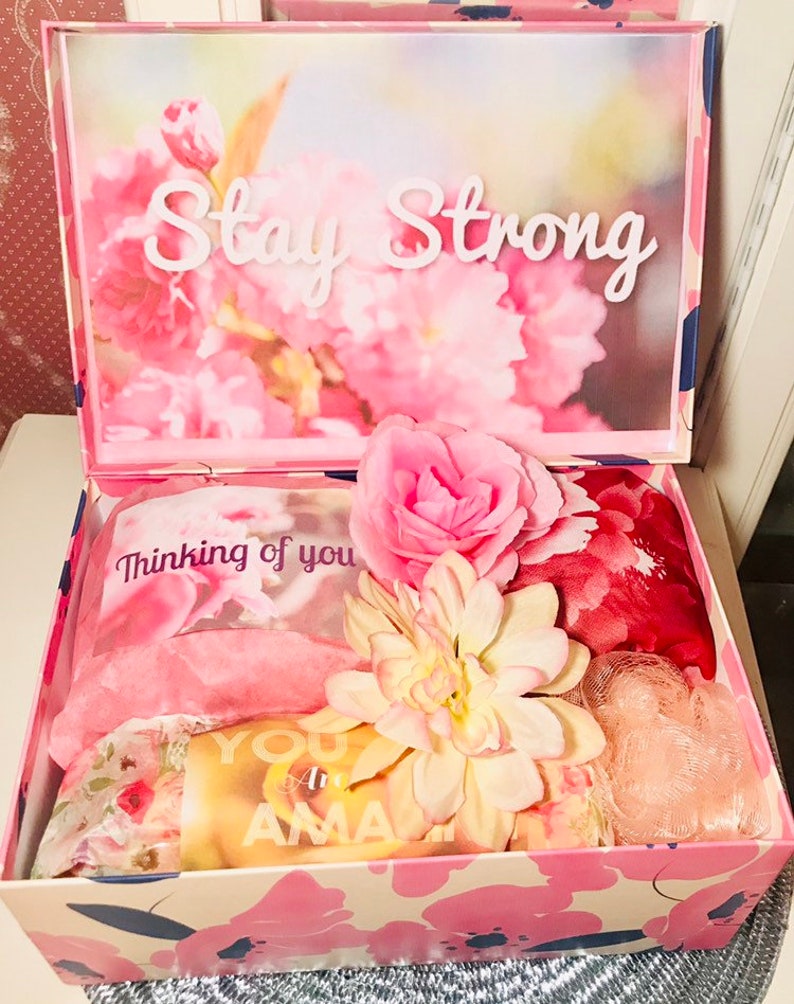 May include: A pink floral gift box with the words "Stay Strong" on the lid. Inside are pink and floral items, including a scarf, a flower, and a card that says "You are Amazing".