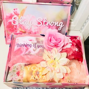 May include: A pink gift box with a floral design and the words "Stay Strong" on the lid. Inside, the box contains pink tissue paper, a card that says "Thinking of you", and a yellow card that says "You are Amazing". Decorative flowers are also included.