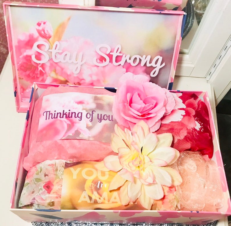 May include: A pink gift box with a floral design and the words "Stay Strong" on the lid. Inside, the box contains pink tissue paper, a card that says "Thinking of you", and a yellow card that says "You are Amazing". Decorative flowers are also included.