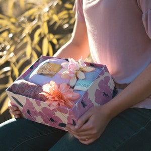 May include: A gift box with a floral design in pink and white, held by a person. The box contains a folded blanket, a scarf, and a tag that says "You Amazing" and "Laugh Love". The box is decorated with pink flowers.