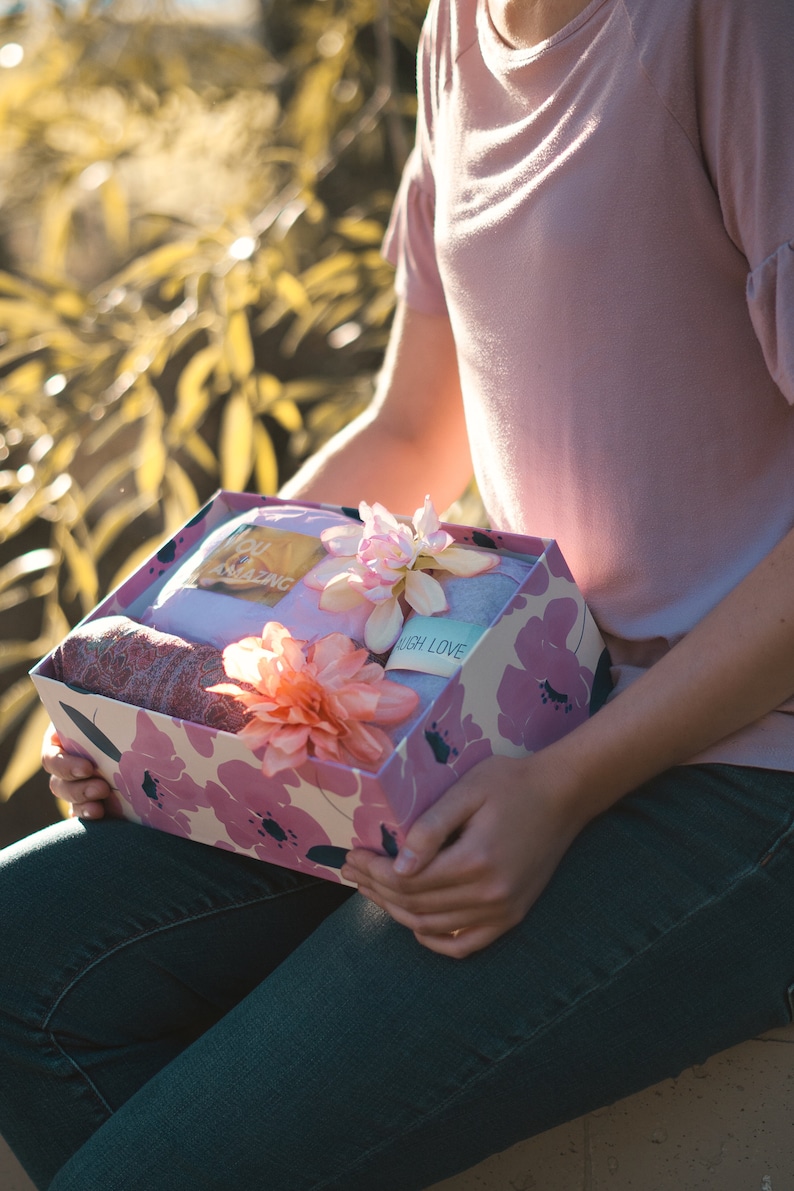 May include: A gift box with a floral design in pink and white, held by a person. The box contains a folded blanket, a scarf, and a tag that says "You Amazing" and "Laugh Love". The box is decorated with pink flowers.
