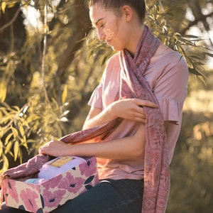 May include: A woman wearing a pink top and jeans holds a floral gift box and a patterned scarf. The scarf is draped around her neck and has a detailed design with fringe. The gift box has a pink floral pattern.