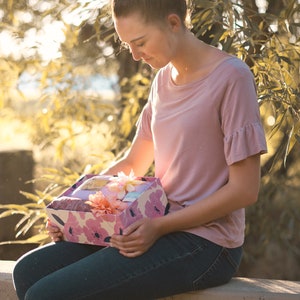 May include: A decorative gift box with a floral pattern in pink and white, held by a person. The box contains a variety of items, including a pink scarf and artificial flowers. The person is wearing a pink top and blue jeans.