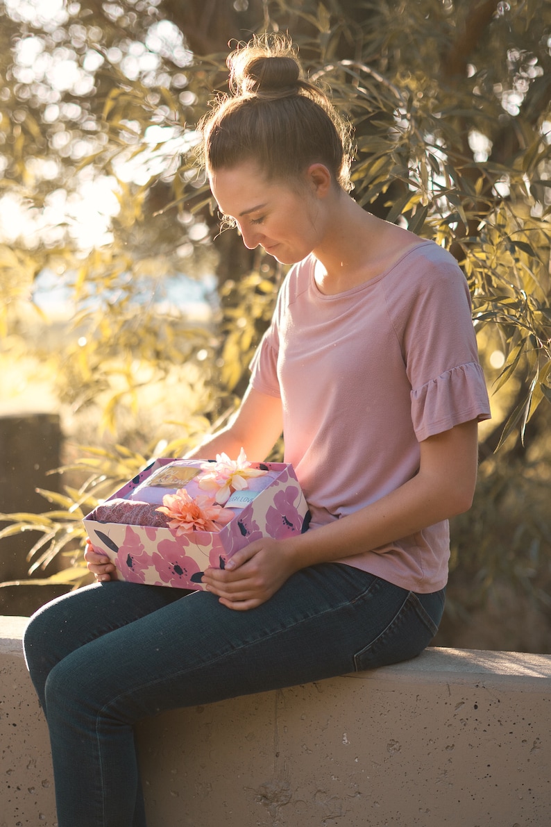 May include: A decorative gift box with a floral pattern in pink and white, held by a person. The box contains a variety of items, including a pink scarf and artificial flowers. The person is wearing a pink top and blue jeans.