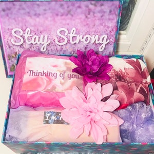 May include: A gift box with a floral design and the words "Stay Strong" on the lid. Inside, a card reads "Thinking of you," alongside a pink flower, a scarf, and other items. The box is decorated with pink and purple accents.