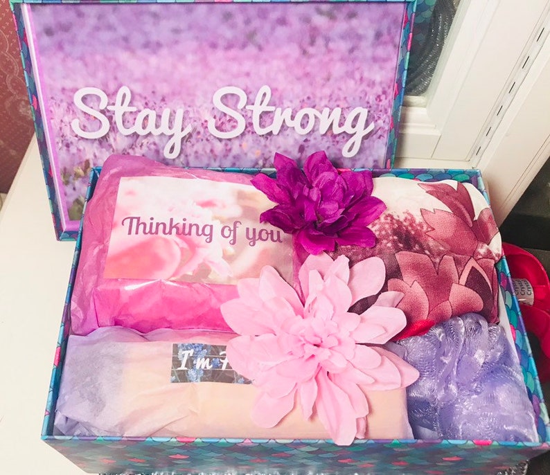 May include: A gift box with a floral design and the words "Stay Strong" on the lid. Inside, a card reads "Thinking of you," alongside a pink flower, a scarf, and other items. The box is decorated with pink and purple accents.