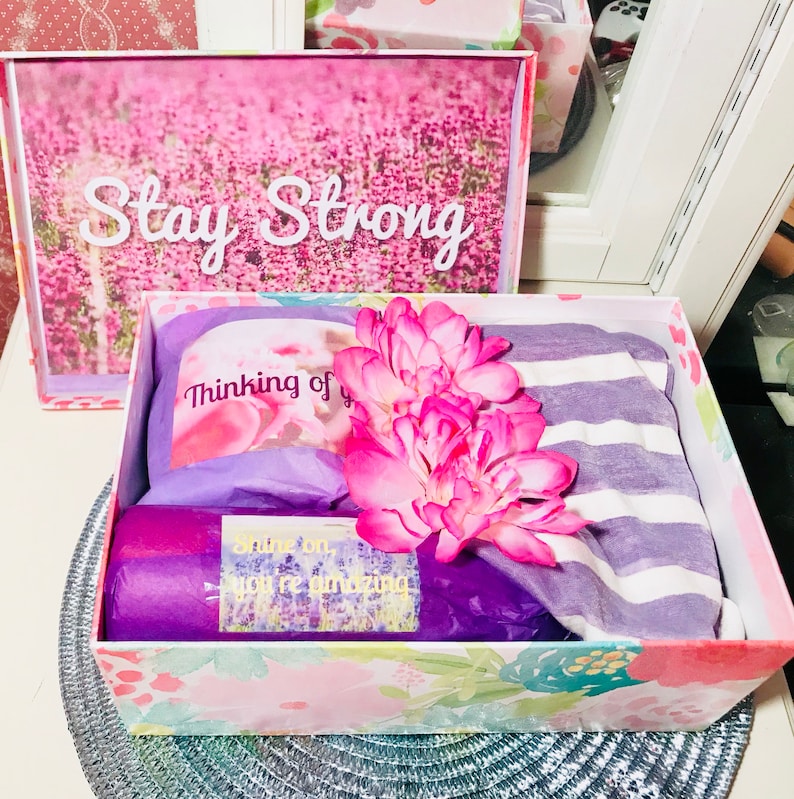 May include: A gift box with a floral design, containing a striped shirt, a card that says "Thinking of you", and a rolled item. A framed picture with the words "Stay Strong" is in the background. A pink flower is on top.