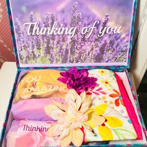 May include: A gift box with a lavender field scene on the lid and the words "Thinking of you." Inside are wrapped items, including a yellow item with "You are Amazing" printed on it, and a floral patterned item. Artificial flowers are on top.