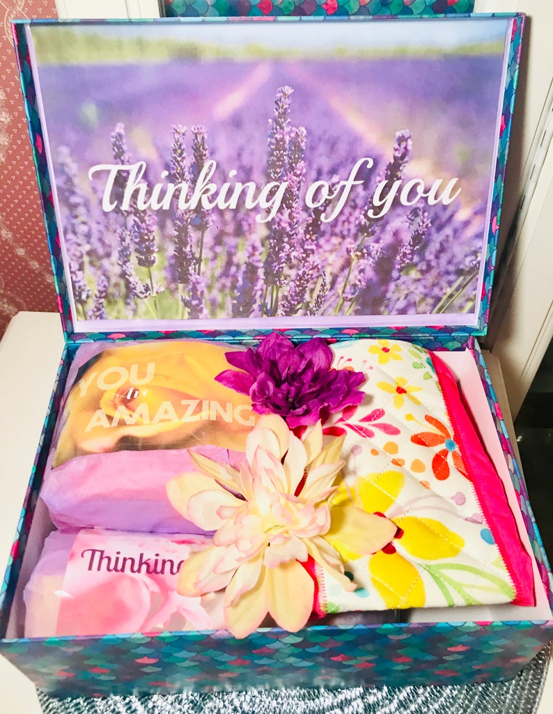 May include: A gift box with a lavender field scene on the lid and the words "Thinking of you." Inside are wrapped items, including a yellow item with "You are Amazing" printed on it, and a floral patterned item. Artificial flowers are on top.