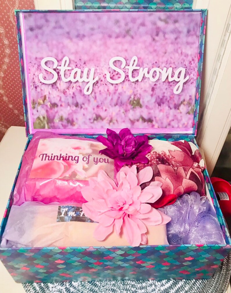 May include: A gift box with a floral design and the words "Stay Strong" on the lid. Inside, the box contains a card that says "Thinking of you", pink tissue paper, and other items. The box is decorated with a mermaid scale pattern.