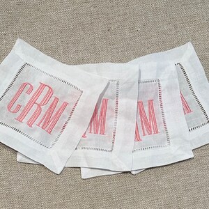 May include: Four white linen cocktail napkins with a square embroidered border and a three-letter monogram in a coral color. The napkins are arranged in a slightly overlapping pattern on a neutral-colored surface.