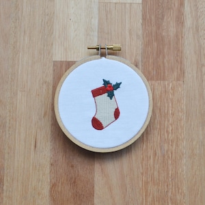 May include: A white embroidery hoop with a red and white Christmas stocking with a green holly sprig on top.