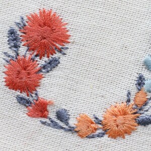May include: A close-up of a white fabric with a floral embroidery design. The embroidery features a wreath of orange, peach, and blue flowers.