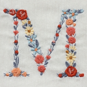 Pode incluir: Letra M bordada com um design floral. A letra &eacute; formada por uma guirlanda de flores azuis, amarelas e laranja.