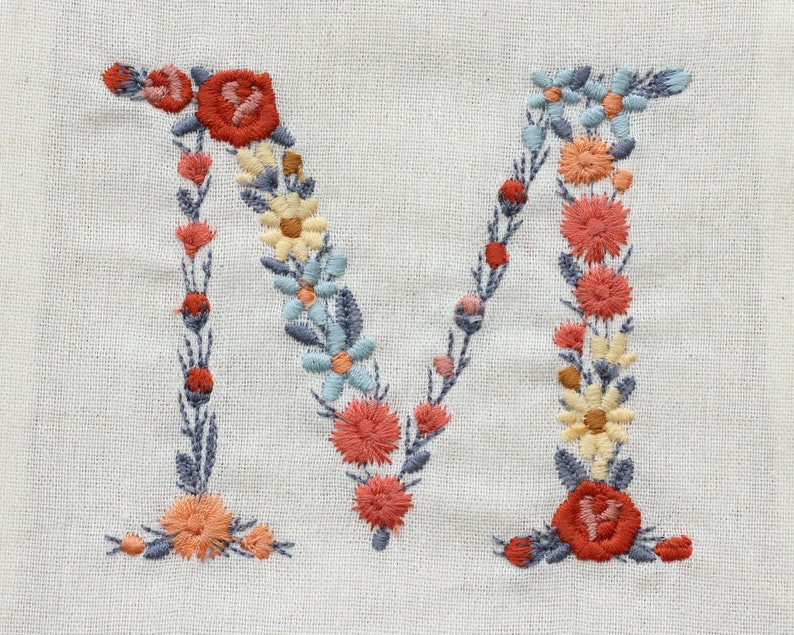 Pode incluir: Letra M bordada com um design floral. A letra &eacute; formada por uma guirlanda de flores azuis, amarelas e laranja.