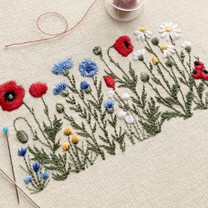 May include: Hand-stitched floral embroidery showing red poppies, blue cornflowers, and white daisies with yellow centres. The design is on beige fabric, with green stems and leaves. Two pins and red thread are also visible.