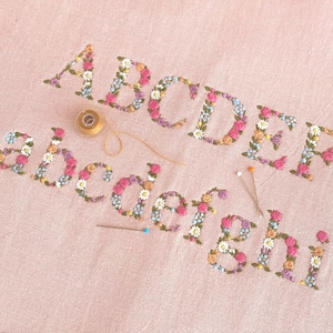 May include: Embroidered uppercase letters A, B, C, D, E and lowercase letters a, b, c, d, e, f, g, h, i on light pink fabric. Each letter is formed by small colourful floral patterns in pink, yellow, blue, and green. A gold spool of thread and two sewing pins are present.