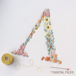 May include: An embroidered letter A on white fabric, featuring a floral design with pink, yellow, blue, and orange thread. A yellow spool of thread and a needle with a red head are positioned at the bottom, with the text DIGITAL FILES.