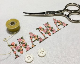 May include: An embroidered piece spelling "MAMA" in floral letters, with pink, peach, and green thread. A spool of yellow thread, small scissors, and two white buttons are arranged on the white fabric. The scissors are approximately 10 cm long.