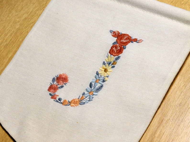 May include: A white fabric banner with the letter "J" embroidered in colourful flowers. The flowers are red, orange, yellow, and blue.