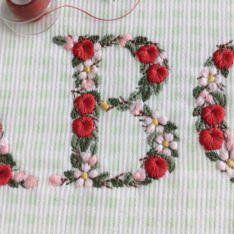 Puede incluir: Primer plano de una tela blanca con las letras "B" y "E" bordadas en rojo, verde y rosa. Las letras est&aacute;n formadas por un patr&oacute;n de peque&ntilde;as flores y bayas.