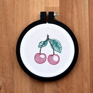May include: Embroidered hoop art featuring two red cherries with green leaves on a white background.