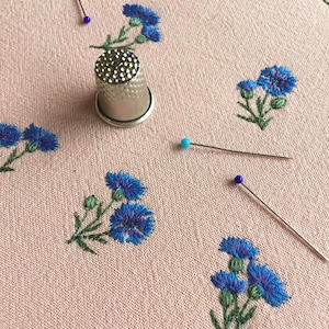 Puede incluir: Una tela de color rosa claro con motivos bordados de acianos azules y tallos verdes. Un dedal de metal plateado se encuentra sobre la tela, acompañado de tres alfileres con cabezas esféricas azules y turquesas dispersos cerca.